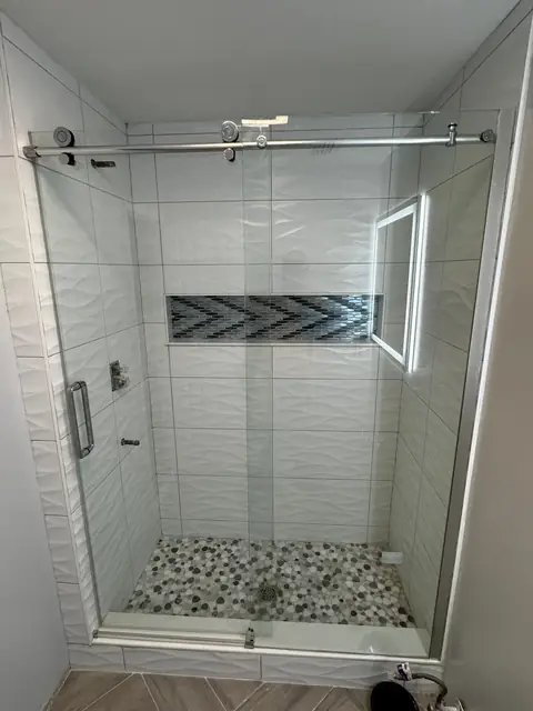 Frameless glass shower door over textured white tile and pebble floor